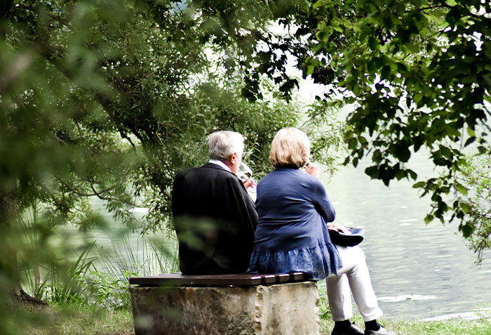 An elderly couple sitting by a lake surrounded by trees, reflecting on deeply disturbing secrets revealed close to them.