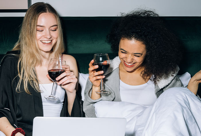 Two women laughing and holding wine glasses while watching something on a laptop revealing disturbing secrets about someone close