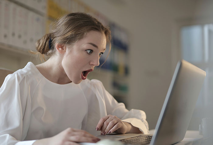 Woman in white blouse looking shocked at laptop screen, revealing disturbing secrets about someone close to her.