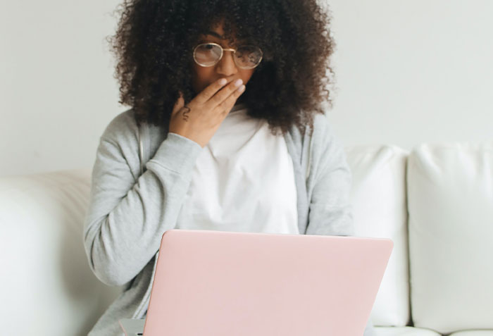Woman with glasses sitting on a couch, looking shocked while uncovering disturbing secrets on her laptop.