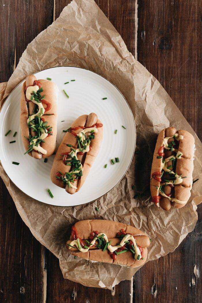 Four hot dogs topped with ketchup, mustard, and chives on a white plate and brown parchment, illustrating things not to buy cheap.
