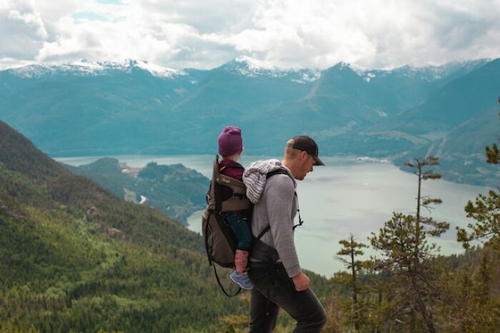 Man hiking with child in backpack overlooking mountain lake and forest, capturing terrifying great outdoors experience.