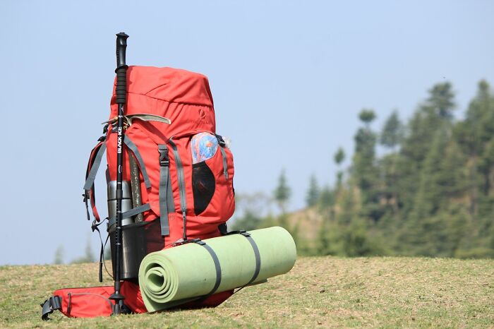 Red hiking backpack with trekking poles and rolled camping mat on grassy hill, representing items not to buy cheap.