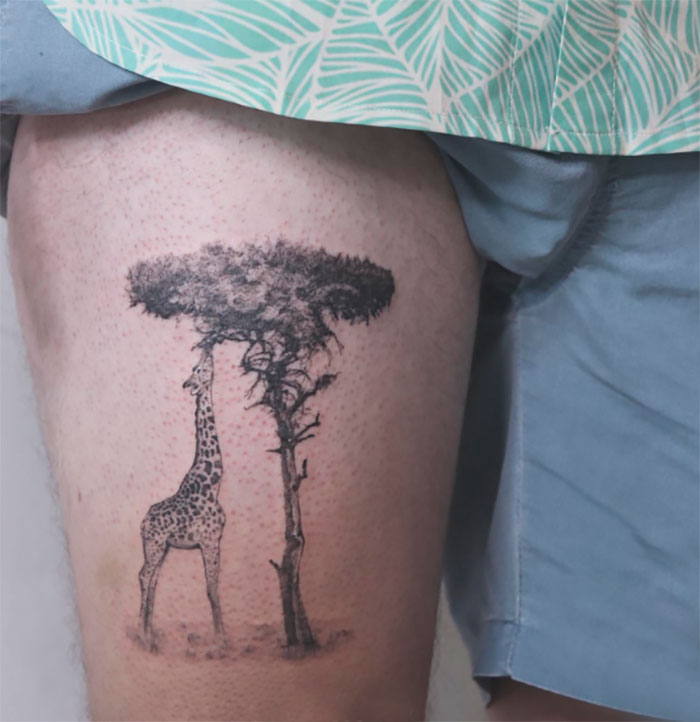 Tattoo of a giraffe standing under a tree on a person's thigh, showcasing nature's wonders and majestic design.
