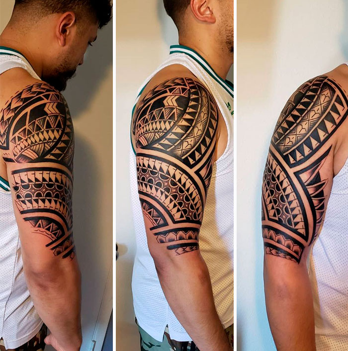 Man with a tribal tattoo sleeve on his upper arm, featuring bold geometric patterns, wearing a white tank top.