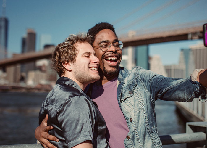 Two college students smiling and taking a selfie outdoors with a city bridge in the background, representing unique college courses.