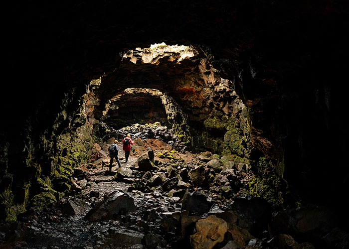 Two hikers exploring a rocky cave tunnel illuminated by natural light, representing unique college courses outdoors.
