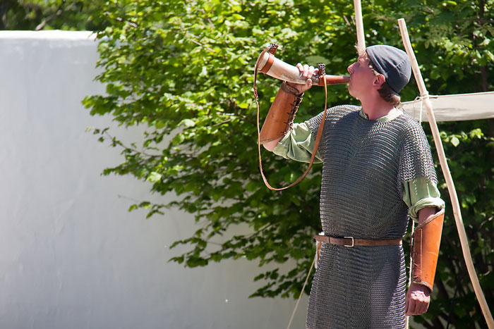 Viking reenactor in chainmail drinking from a horn outdoors with green trees in the background during a sunny day