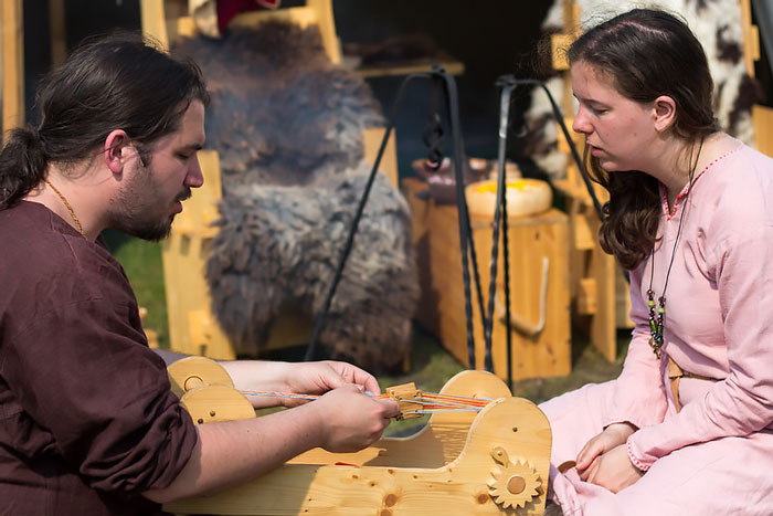 Two people dressed as Vikings demonstrating a traditional Viking weaving tool at an outdoor historical event.