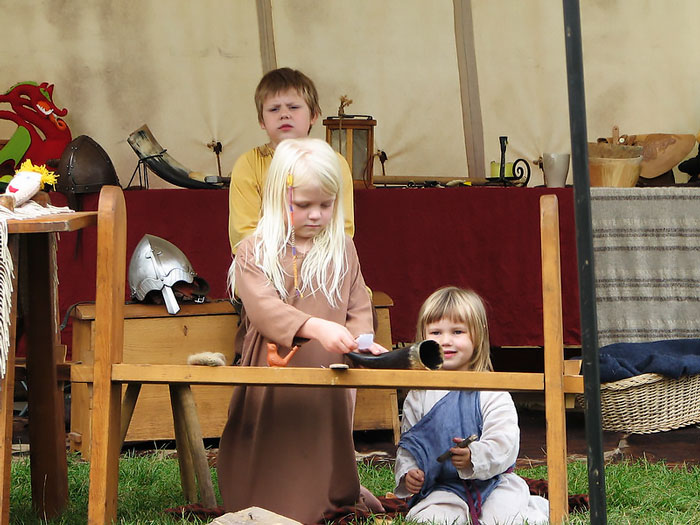 Three children dressed in Viking-style clothing playing with Viking artifacts at a historical reenactment event.