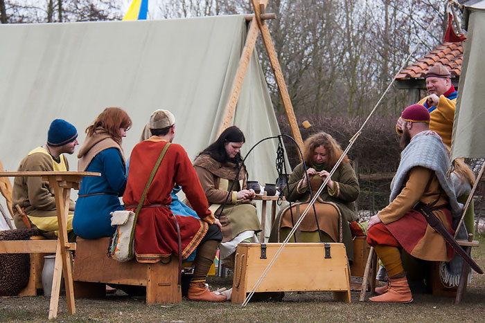 Group of people dressed in historical Viking attire sitting and interacting around a campfire at a reenactment event.