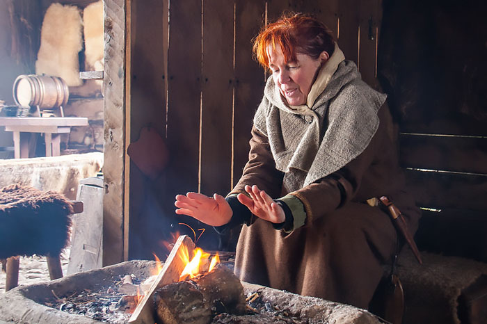 Woman dressed in Viking clothing warming her hands by a fire inside a rustic wooden Viking shelter.