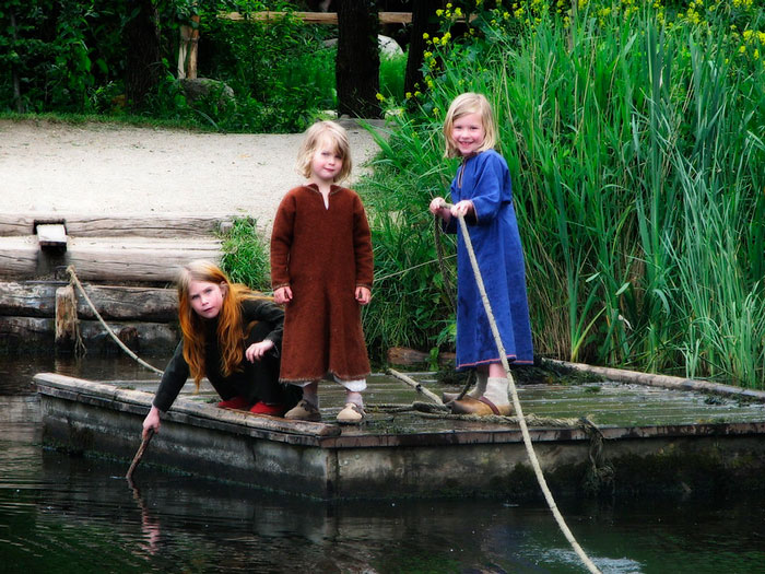 Three children dressed in Viking-style clothing on a wooden raft near a grassy riverbank exploring Viking life.