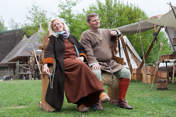Two people dressed in traditional Viking clothing sitting outdoors with rustic Viking-style buildings in the background.