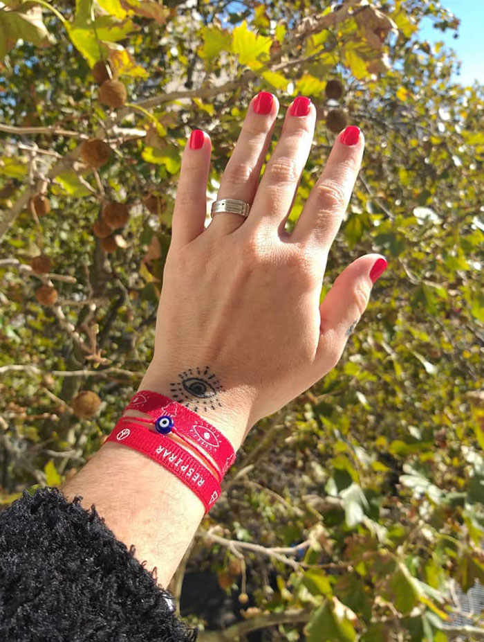 A wrist tattoo of an eye design is shown, with red bracelets and a ring on the hand, set against a leafy background.