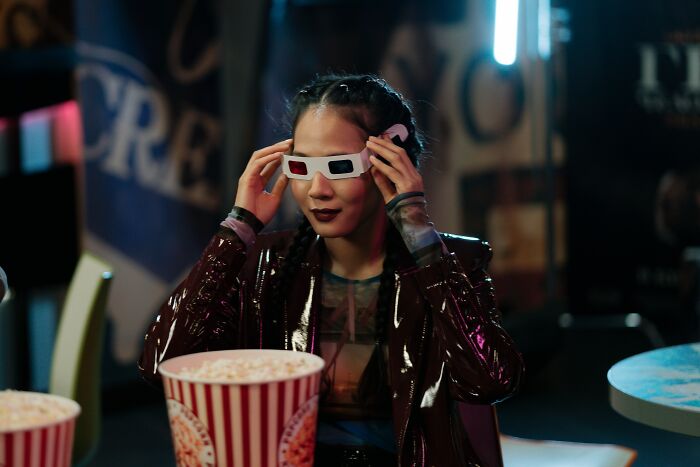 Young woman wearing 3D glasses and a shiny jacket, holding popcorn, capturing a moment of human stupidity humor.