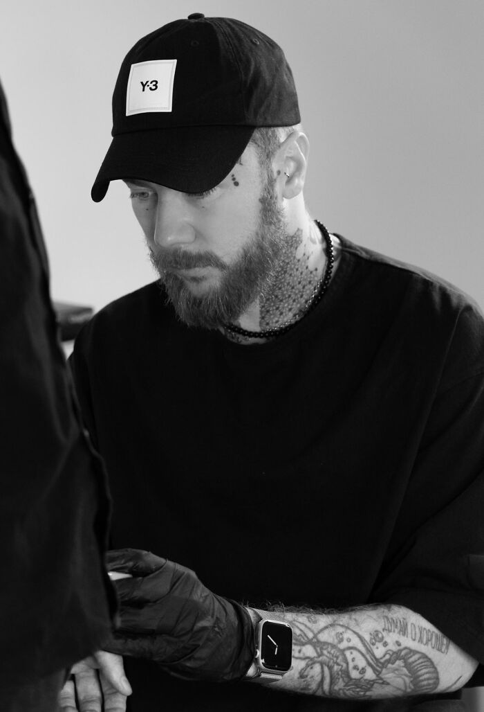 Tattoo artist focused on intricate geometric design, wearing a cap and gloves, showcasing detailed arm tattoos.