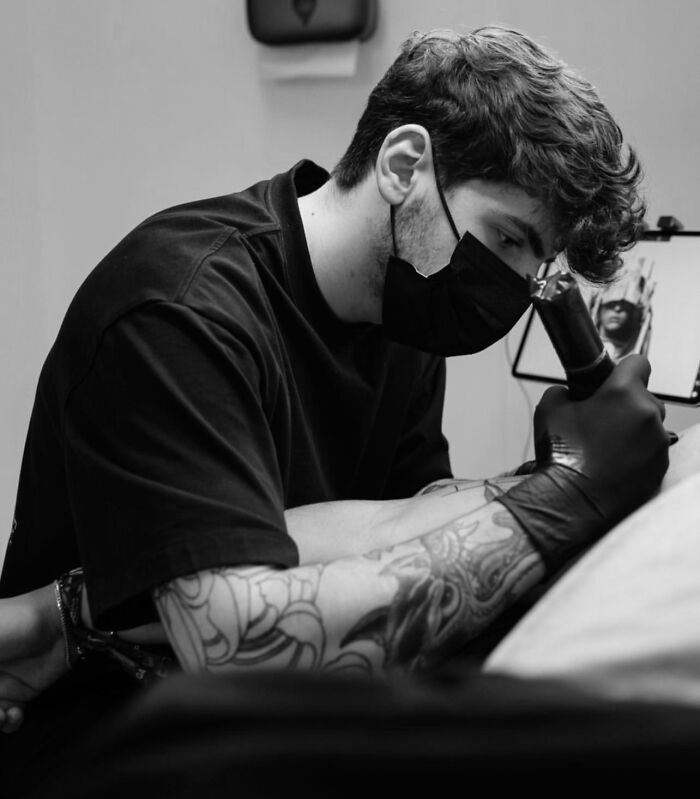 Tattoo artist creating geometric design; black mask, gloves, and detailed arm tattoos visible.