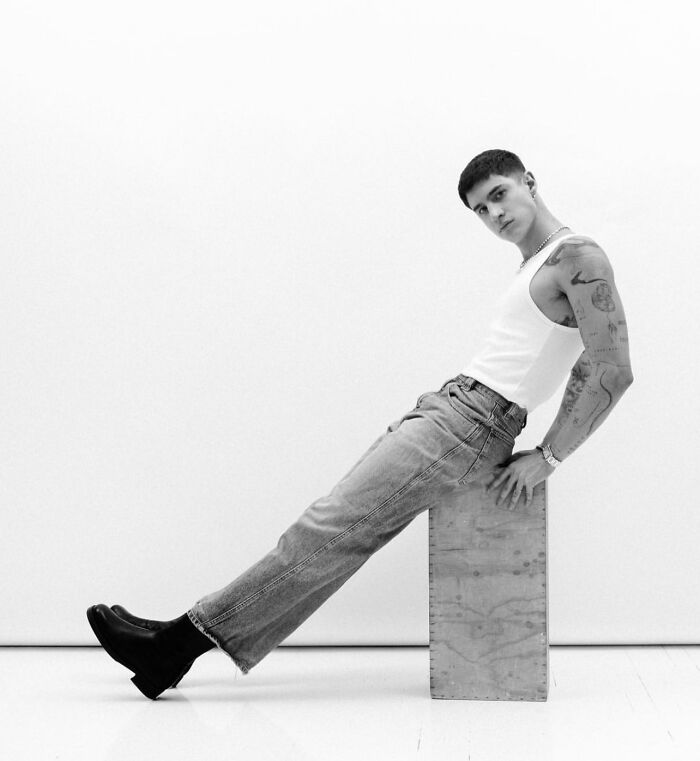 Person in a white tank top and jeans, showcasing geometric tattoos in a minimalist setting.