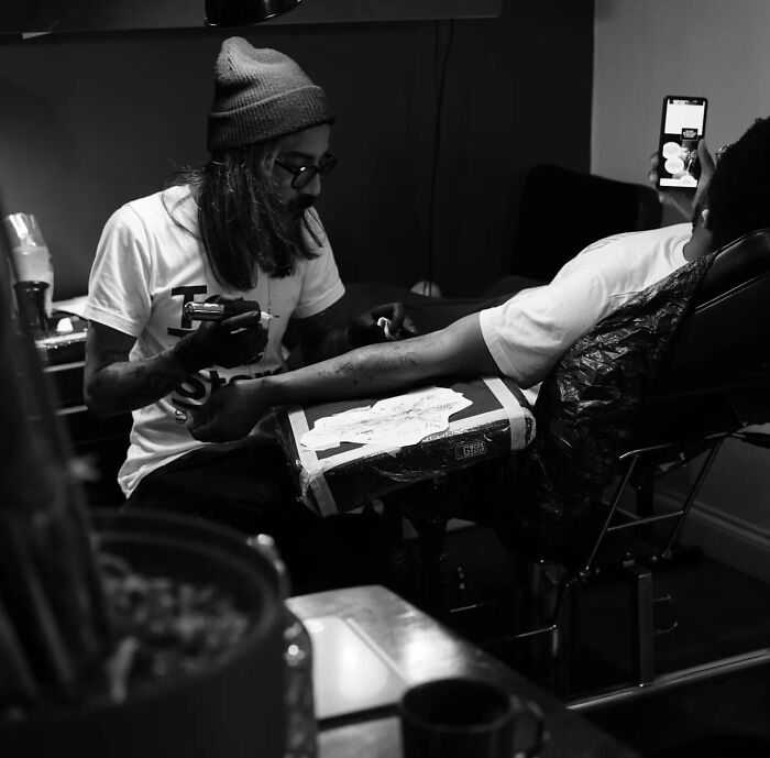 Tattoo artist working on a geometric tattoo, wearing a beanie and glasses, in a dimly lit studio.