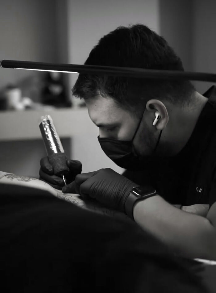 Tattoo artist focused on creating a geometric tattoo, wearing a mask and gloves, holding a tattoo machine.