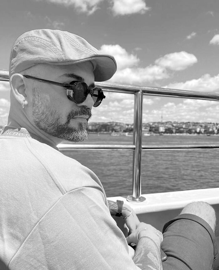 Man with beard and sunglasses on a boat, showcasing geometric tattoo style, ocean and cityscape in the background.