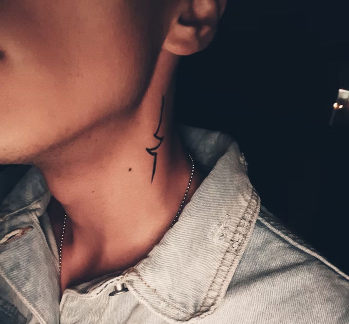 Minimalist tattoo on a man's neck, wearing a denim jacket, in dim lighting.