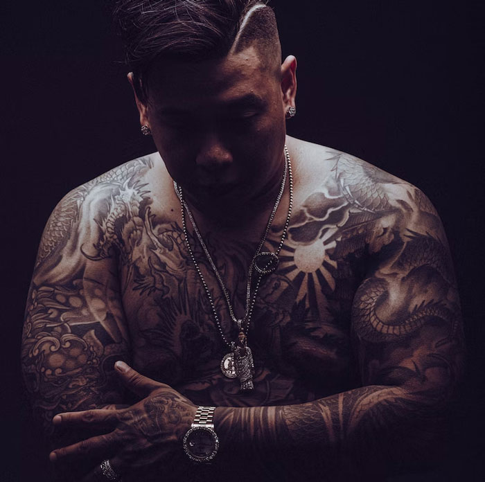 Man with tattoos covering arms and chest, standing with arms crossed, wearing jewelry, in a dimly lit setting.