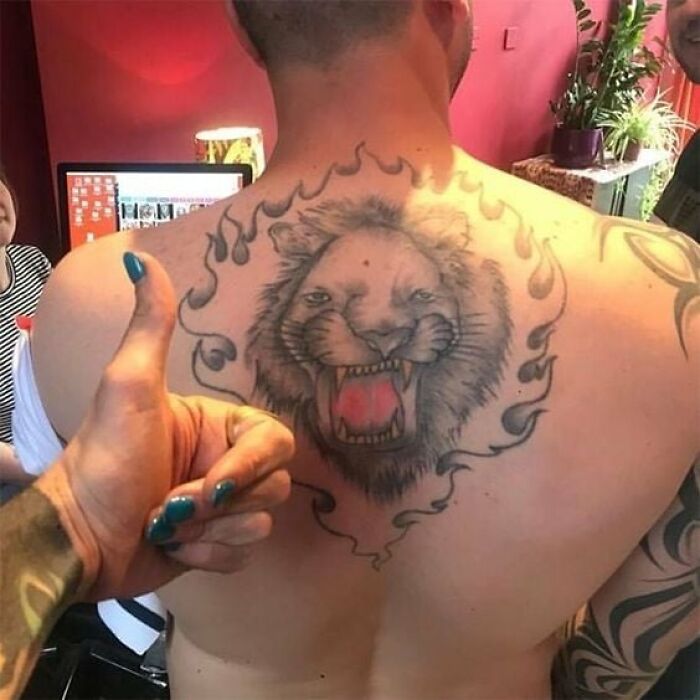 Back tattoo of a lion surrounded by flames, with a person giving a thumbs-up in the foreground, illustrating tattoo fails.