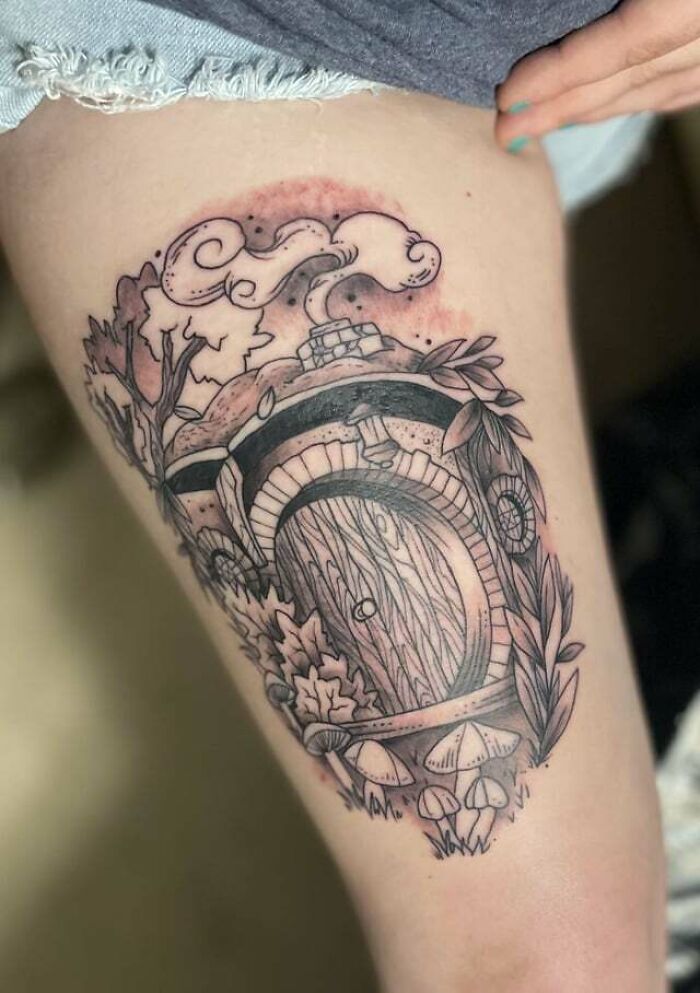 Tattoo of a hobbit door from Lord of the Rings, detailed with leaves and mushrooms on a person's thigh.