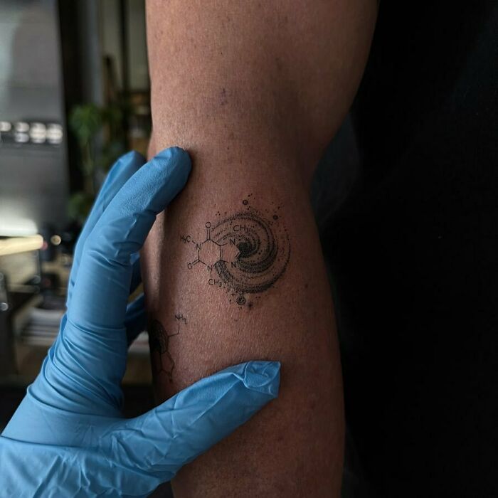 Science tattoo featuring a molecular structure and galaxy design on an arm, with blue-gloved hand examining it.