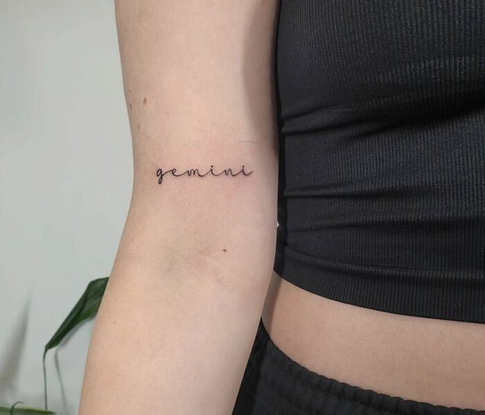 Gemini zodiac tattoo in minimalist script on a person's arm wearing a black top.