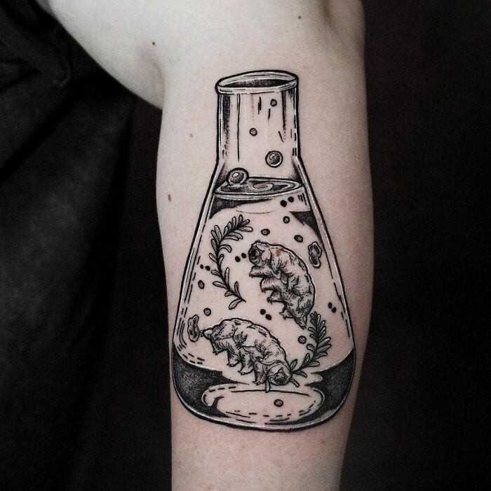 Science tattoo of a beaker with tardigrades and foliage, inked on forearm.