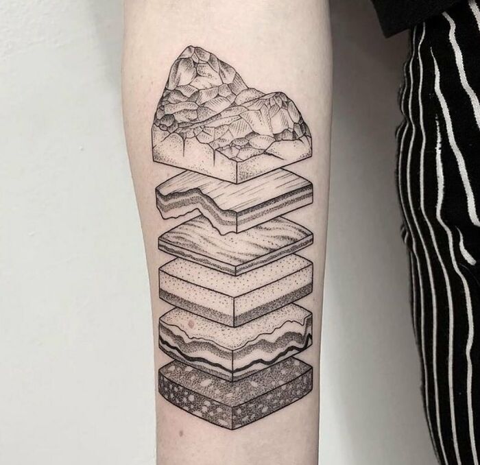 Geology-themed science tattoo on forearm, depicting layered rock formations in a detailed black ink design.