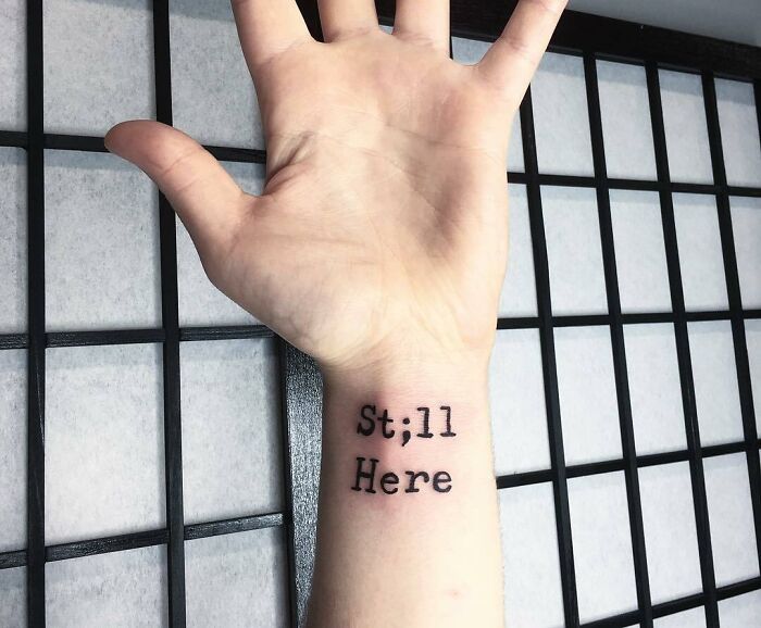 Tattoo on wrist reads "St;ll Here" symbolizing mental health resilience.