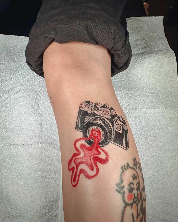Abstract tattoo of a camera and red splash on a forearm, showcasing artistry and self-expression.