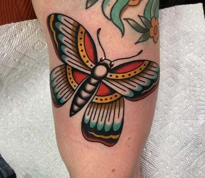 Colorful butterfly tattoo with intricate patterns on an arm, showcasing vibrant red, teal, and gold hues.