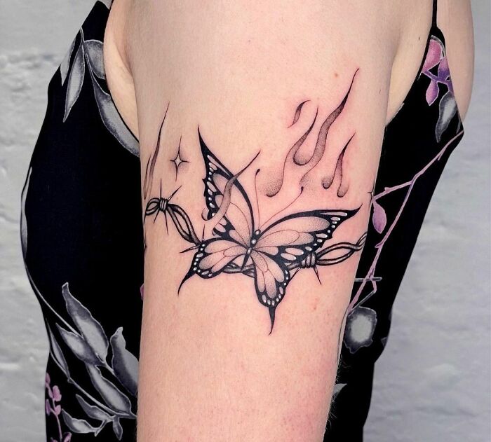 Butterfly tattoo with flames and barbed wire on upper arm, showcasing intricate design and bold black lines.
