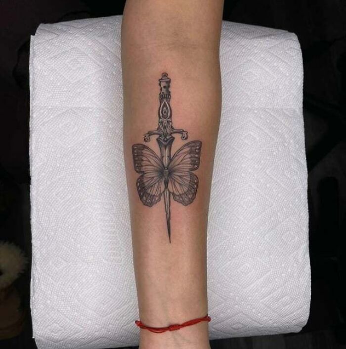 Butterfly tattoo with dagger design on forearm, showcasing intricate black ink details.