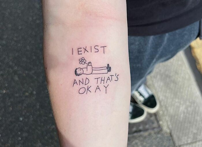 Mental health tattoo on forearm with text "I exist and that's okay," featuring a small figure holding a flower.