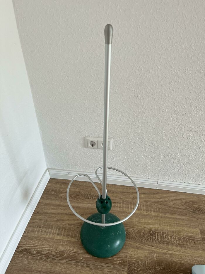 Tall mysterious object with a green base and looped metal wire, standing on wooden floor against a white wall surface.