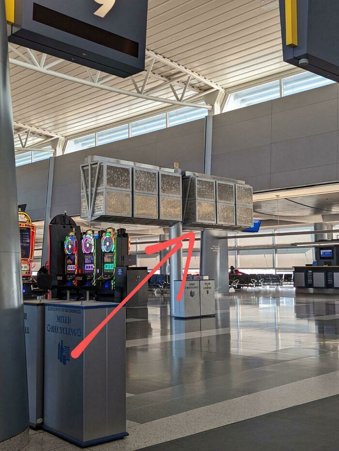 Mysterious object hanging from ceiling in airport terminal near slot machines and recycling bins.