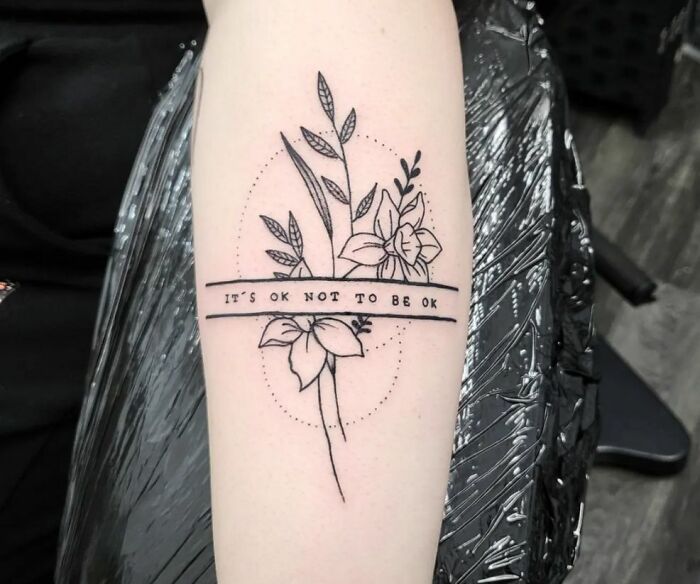 Floral mental health tattoo with text "It's OK not to be OK" on arm, symbolizing healing and self-acceptance.