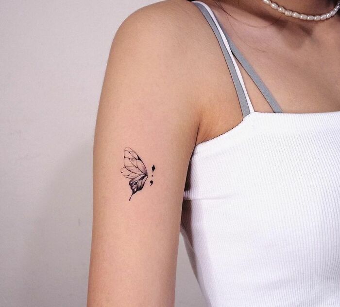Butterfly and semicolon tattoo on woman's upper arm, symbolizing mental health and healing.