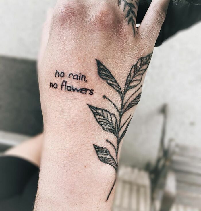 Hand with a mental health tattoo saying "no rain, no flowers," alongside a leafy vine design.