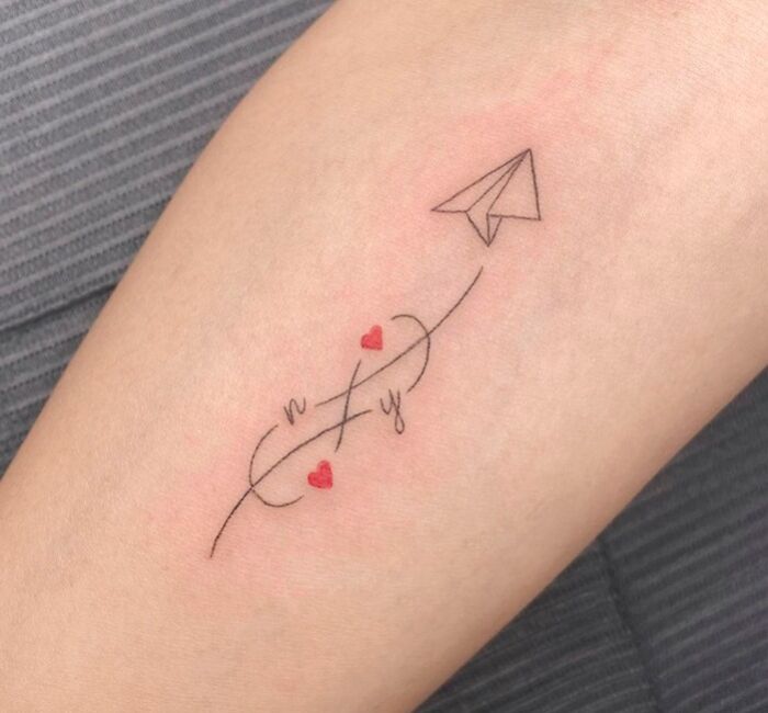 Infinity tattoo with paper airplane and red hearts on arm, symbolizing eternal creativity.