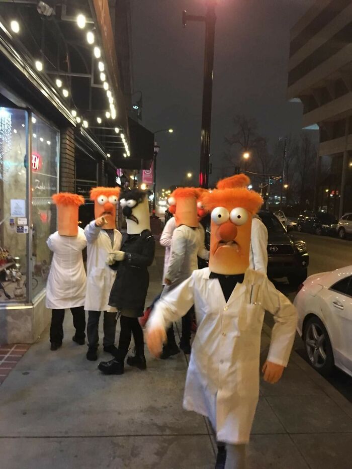 Group of people wearing weird costumes with oversized orange heads on a city sidewalk at night, showing bizarre and weird pics.