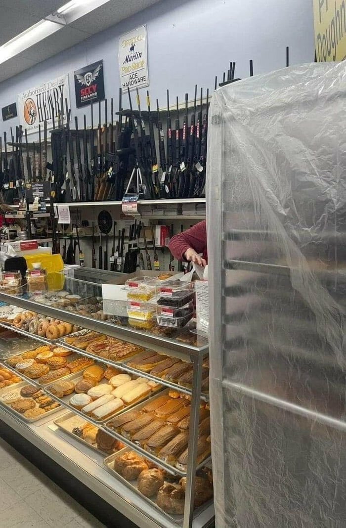 Bakery display case filled with pastries next to a large rack of rifles in a unique weirdest pics setting.