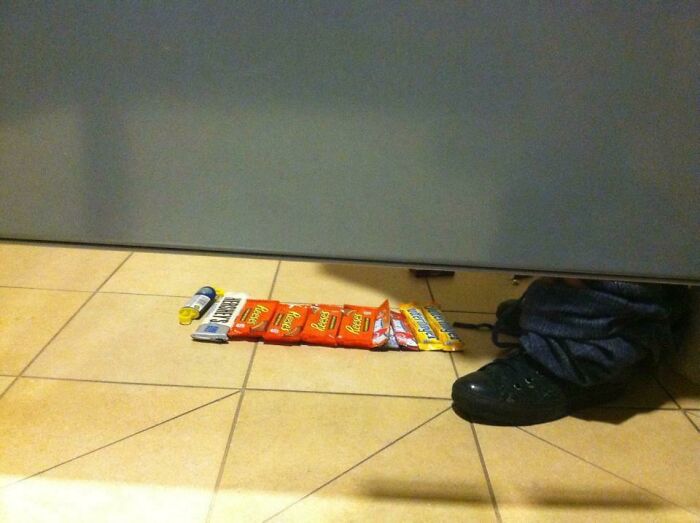 Shoes and pants visible under a restroom stall with various candy bars and chapstick lined up on the floor in a weirdest pics scene.