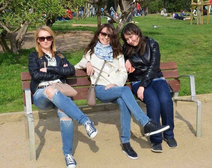 Three women sitting on a park bench with crossed legs, enjoying a sunny day and casual outdoor setting.